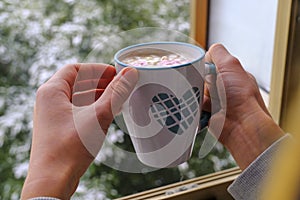 Hands holding mug the blue heart with hot chocolate with pink marshmallows across winter landscape in open window. Winter vacation