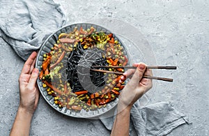 Hands holding a plate of black spaghetti or pasta with grilled vegetables, broccoli, corn, green peas in plate on light gray