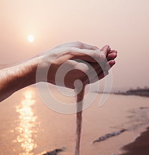 Hands holding and spilling sand with beach at sunset in the background