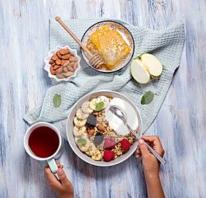 Hands holding spoon of healthy breakfast with granola, berries and yogurt and a cup of tea