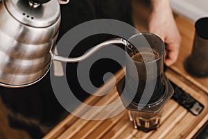 Hands holding steel kettle and aeropress, glass cup, scales, coffee beans on wooden table. Professional barista pouring hot water