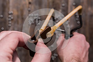 Hands holding a stretched slingshot, ready for shot with a metal ball, on the background of an old wooden fence