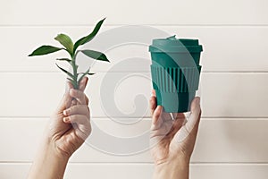 Hands holding stylish reusable eco coffee cup and green bamboo leaves on white wooden background. Zero waste. Green Cup from