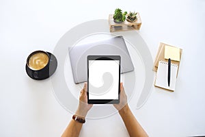 Hands holding a tablet with blank white screen and laptop computer on the table in office