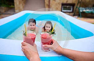 Hands holding two glasses with freshly squeezed fruit juice against blurred background of children swimming in the pool