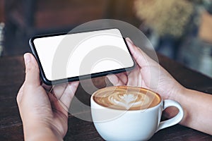 Hands holding and using a black mobile phone with blank screen horizontally for watching with coffee cup on wooden table