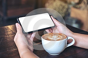 Hands holding and using a black mobile phone with blank screen horizontally for watching with coffee cup on wooden table