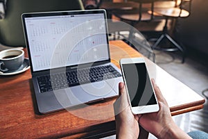 Hands holding and using mockup mobile phone with blank black screen and laptop on wooden table