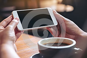 Hands holding and using a white mobile phone with blank black screen horizontally for watching with coffee cup on wooden table