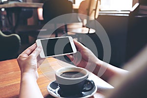 Hands holding and using a white mobile phone with blank black screen horizontally for watching with coffee cup on wooden table