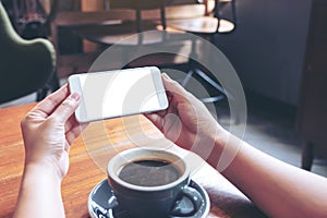 Hands holding and using a white mobile phone with blank screen horizontally for watching with coffee cup on wooden table