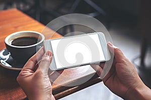 Hands holding and using a white mobile phone with blank screen horizontally for watching with coffee cup on wooden table