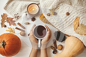 Hands holding warm cup of tea on background of autumn leaves, pumpkin, cozy sweaters, burning candle on white wood. Hello autumn