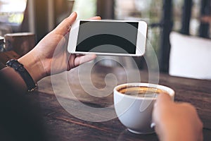 Hands holding white mobile phone with blank black desktop screen and a coffee cup on wooden table in vintage cafe