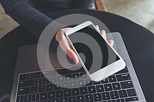 Hands holding white mobile phone with blank black desktop screen and laptop on table in cafe