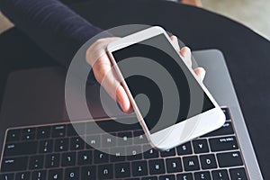 Hands holding white mobile phone with blank black desktop screen and laptop on table in cafe