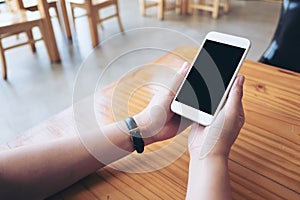 Hands holding white mobile phone with blank black screen on wooden table in cafe