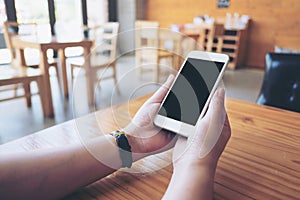 Hands holding white mobile phone with blank black screen on wooden table in cafe