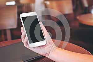 Hands holding white mobile phone with blank desktop screen with laptop computer on the table