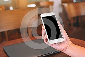 Hands holding white mobile phone with blank desktop screen with laptop computer on the table