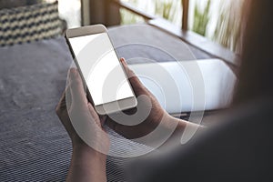 Hands holding white mobile phone with blank desktop screen and laptop on table in cafe