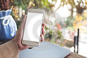 Hands holding white mobile phone with blank desktop screen with laptop on table in cafe