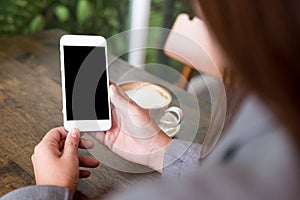 Hands holding white mobile phone with blank white screen with hot coffee cup on wooden table in cafe