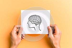 Hands holding white paper with human head and brain on yellow background. Mental health and positive thinking concept.