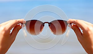 Hands holding women`s sunglasses on the background of the blue sea and the sun.