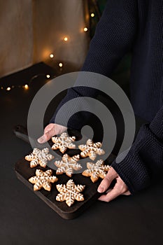 Hands holding wooden board with snowflake gingerbread cookie decorated with sugar icing, on black table background