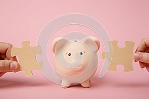 Hands holding wooden jigsaw puzzle pieces near piggy money bank on pastel pink wall background. Money