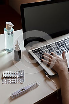 Hands on the keyboards with pile of medical face masks, sanitizer, headphones, nasal drops, pills nearby.  workplace at ho