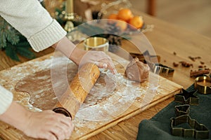 Hands kneading gingerbread dough with rolling pin, cooking spices, festive decorations on rustic table. Woman making christmas