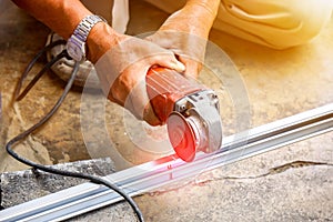 Hands of laborer holding electric angle grinder working cutting aluminium lumber at construction site with sun flare