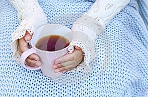 Hands of little girl in cozy hand warmers fingerless gloves holding cup of tea on her knees wrapped in warm knitted plaid