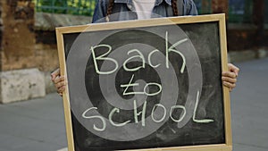 Hands of Little girl standing holding and raising schoolboard with text back to school on the street near school. Close