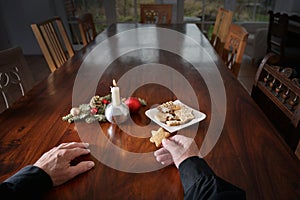 Hands of a lonely old man with a cookie sitting at a big family table with candle, Christmas decoration and empty chairs, holidays