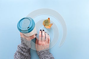 Hands in long sleeve knitted sweater, holding bamboo reusable cup and autumn leaf, overhead on blue background