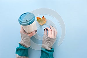 Hands in long sleeve knitted sweater, holding bamboo reusable cup and autumn leaf, overhead on blue background