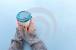 Hands in long sleeve knitted sweater, holding bamboo reusable cup with lid on, overhead on blue background