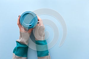 Hands in long sleeve knitted sweater, holding bamboo reusable cup with lid on, overhead on blue background