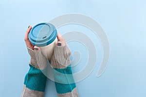 Hands in long sleeve knitted sweater, holding bamboo reusable cup with lid on, overhead on blue background