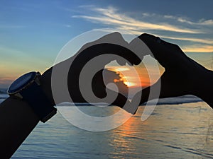 Hands making love sign. Fingers with heart gesture. Sunset in hand on blue beach landscape background. Love hope faith concept. photo