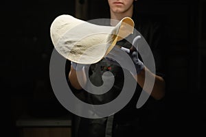 Hands of a male chef with thin round pizza dough in the kitchen on a dark background. Pizzeria concept