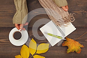 Hands of man with a cup of espresso and scarf at wooden table with notebook and pen and autumn leaves