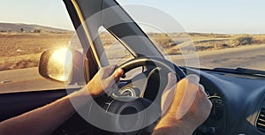 Hands of a man driver on car steering wheel. Dashboard. Windshield and side window view. Arid countryside landscape along the road