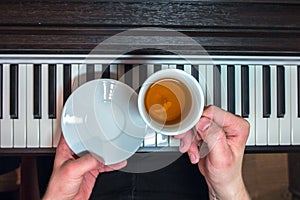 Hands of a man holding a white saucer and a cup of tea on the background of the piano keyboard.