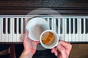 Hands of a man holding a white saucer and a cup of tea on the background of the piano keyboard.