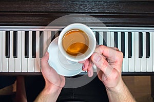 Hands of a man holding a white saucer and a cup of tea on the background of the piano keyboard.