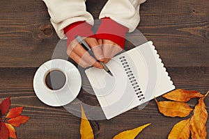 Hands of man with a pen and notebook at a wooden table with cup of espresso and autumn leaves